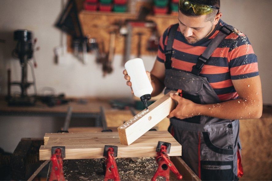 Άνδρας μέσα σε εργαστήριο με σαλοπέτα φόρμα εργασίας βάζει κόλλα σε ένα κομμάτι ξύλο.